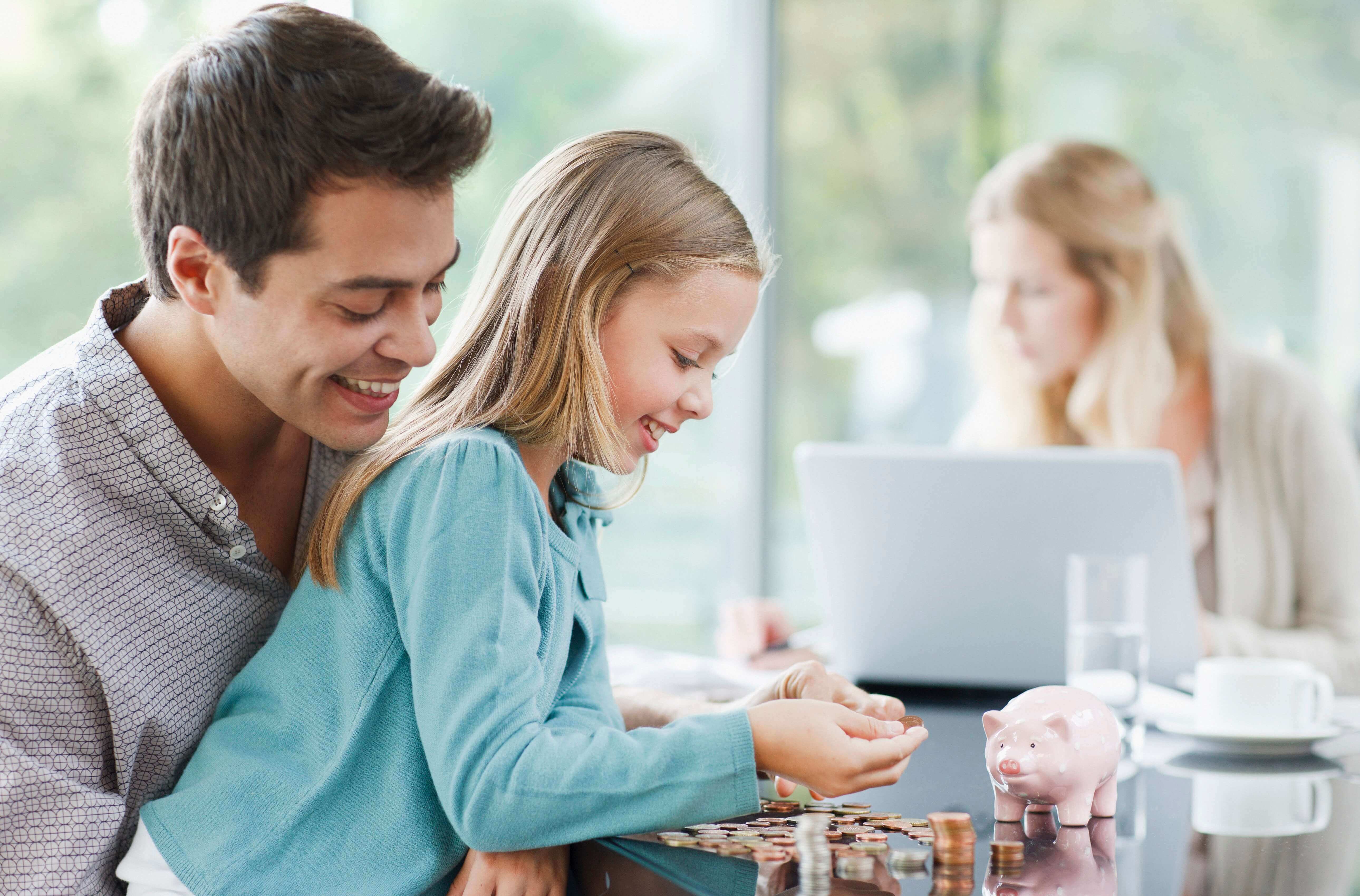 Man and Child Counting Coins next to Piggy Bank, Woman in the Background on a Laptop